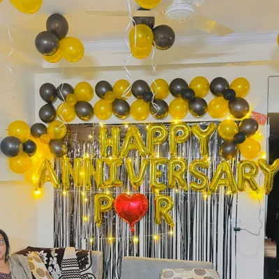 A 'Happy Anniversary' decoration in a living room, using black and gold balloons, a shimmer curtain, and fairy lights.