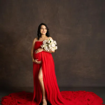 A stunning solo portrait of a mother-to-be in a vibrant red strapless gown with a long train. She holds a bouquet of white flowers, looking off to the side with a gentle smile against a classic brown backdrop.