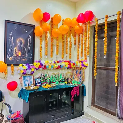 A home pooja corner decorated for a celebration. I used hanging marigold garlands and a line of colorful artificial flowers above the deities, along with some balloons for a festive touch.