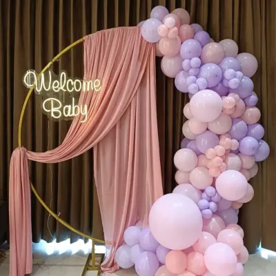 An elegant 'Welcome Baby' setup featuring a gold ring frame draped with soft pink fabric. A pastel pink and purple balloon arch and a warm neon sign complete this beautiful arrangement for a baby shower.