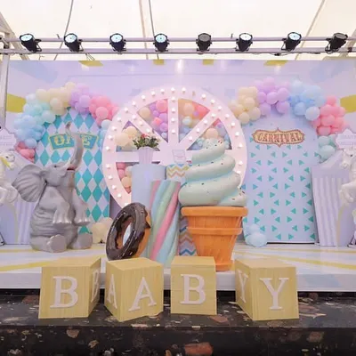 A dreamy pastel carnival stage setup for a first birthday. The scene includes a lit Ferris wheel, giant ice cream cone and donut props, and adorable animal figures, all in front of a custom-designed geometric backdrop.