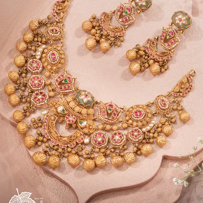 A legacy that adorns generations. This heritage bridal necklace, with its intricate patterns, ruby and emerald-hued stones, and clusters of golden ghungroos, is handcrafted to tell your story in gold and grace. It is an heirloom in the making.