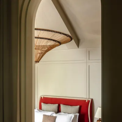 An arched doorway frames the master bedroom in our 'Summer Haus' project, creating a sense of theatrical entry. The view reveals a bold red headboard and a striking rattan light fixture, hinting at the eclectic and cosmopolitan design within.