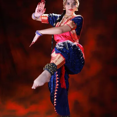 A dynamic full-body shot of a Bharatanatyam dancer in mid-pose. The dramatic red background and studio lighting make the dancer's form and colorful costume stand out.