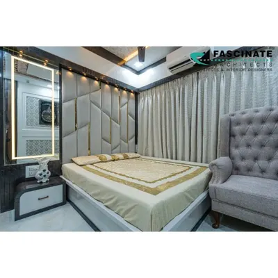 A luxurious master bedroom in Jogeshwari featuring a floor-to-ceiling upholstered headboard with geometric gold inlay. The backlit vanity mirror and plush wingback chair add to the room's sophisticated and comfortable feel.