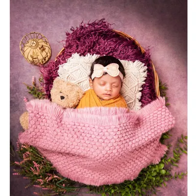 A perfect little bundle sleeping soundly in a basket. The baby is wrapped in a warm mustard-colored cloth and tucked in with a pink blanket, accompanied by a tiny teddy bear.