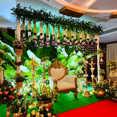 A lush green Seemantham stage featuring a canopy of leaves, hanging jasmine and purple flower garlands, and a traditional white and gold chair for the ceremony.