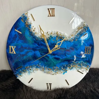 A close-up of a geode-inspired wall clock. The deep blue resin flows beautifully into the white, with shimmering gold crystals creating a natural, luxurious divide. The gold Roman numerals complete this elegant timepiece.