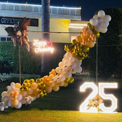 An elegant outdoor setup for a 25th anniversary party. This features a diagonal balloon garland, a custom neon sign, dried floral arrangements, and a large illuminated '25'.