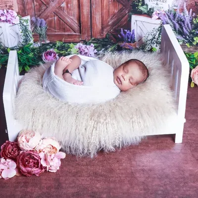 A classic newborn pose with the baby swaddled and sleeping peacefully in a miniature white bed. The rustic floral backdrop adds a touch of warmth and charm.