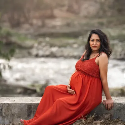 An expecting mother in a flowing red dress sits peacefully by a flowing stream. This outdoor portrait combines the raw beauty of nature with a serene, contemplative mood.