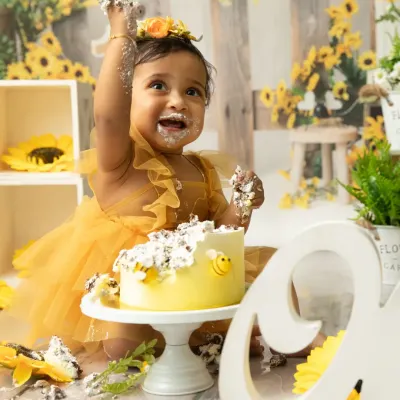 A sunshine-filled first birthday with a sunflower theme. This little one is having the time of her life, smashing her cake with pure delight in a bright and cheerful studio setup.