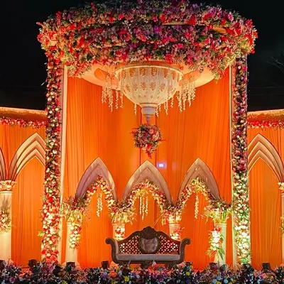 A magnificent wedding stage with a royal palace design, featuring grand arches, columns, and a huge floral dome, all in a warm orange and gold palette.