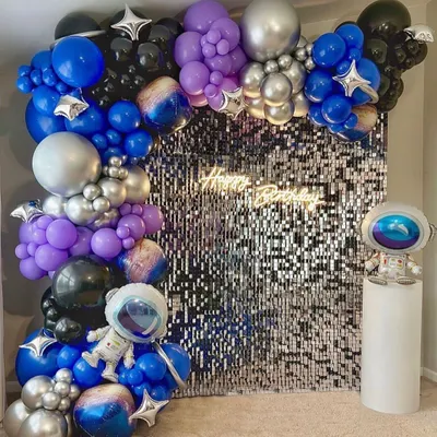 An out-of-this-world space theme birthday party. This setup features a silver shimmer wall, a planet-themed balloon garland in blue, purple, and silver, and astronaut props.