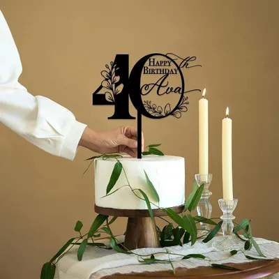 A hand places a custom "Happy 40th Birthday Ava" topper onto a simple white cake. This intricate black acrylic design features floral details for an elegant and personalized touch.