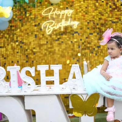 A complete setup for a first birthday photoshoot, featuring a sparkly gold backdrop, a light-up name sign, and pastel balloons. The birthday girl is dressed in a beautiful party dress to complete the look.