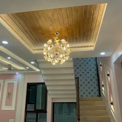 A luxurious villa interior featuring a stunning crystal chandelier hanging from a wooden false ceiling panel. The multi-level ceiling design and wall molding add depth and grandeur to the space.