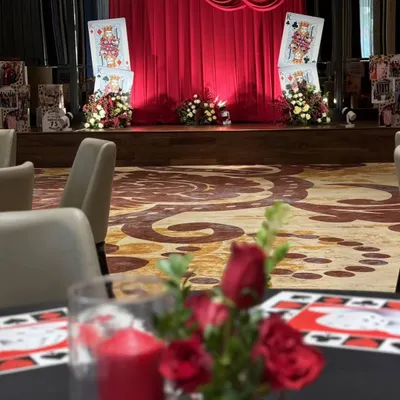 A view from a guest's table at our casino-themed event. The red rose centerpiece in the foreground complements the dramatic red draping of the main stage in the background, creating a cohesive and luxurious atmosphere throughout the room.