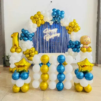 A classic first birthday setup with a blue, yellow, and white balloon arch. This decoration includes a "Happy Birthday" neon sign against a blue fringe curtain and a large number '1' foil balloon.
