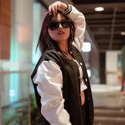 A classic streetwear look featuring a varsity jacket and sunglasses. The shallow depth of field and the model's confident pose create a cool, urban vibe perfect for a youth-focused fashion brand.