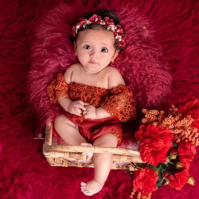A striking portrait of a three-month-old baby girl. Dressed in a lacy red outfit with a matching floral headband, she sits in a small basket against a rich, textured red background.