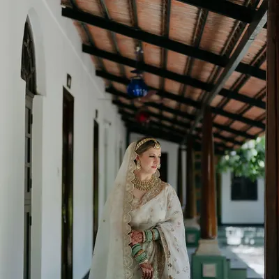A bride in a stunning white saree stands in a sunlit corridor, the repeating pillars creating a beautiful, elegant frame for her portrait.