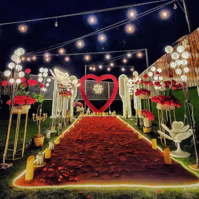 A nighttime view of a spectacular proposal setup. The combination of a red rose petal aisle, a glowing heart arch, and dozens of LED candles creates an intensely romantic and picture-perfect scene against the dark sky.