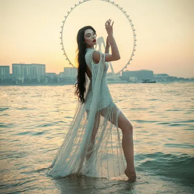 A model strikes an artistic pose in the shallow waters of a Dubai beach, her flowing white dress echoing the shape of the Ain Dubai ferris wheel at sunset.