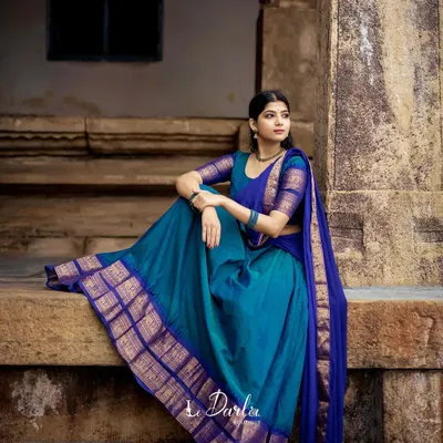 A moment of quiet contemplation in a beautiful peacock blue and royal blue half-saree. The rich silk fabric and its golden border are highlighted by the serene temple setting.