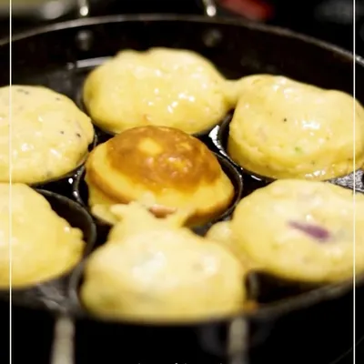 A close-up of Muttai Kuzhi Paniyaram sizzling in a traditional cast-iron pan. These savory Chettinad-style egg and rice batter puffs are cooked until golden brown and fluffy.