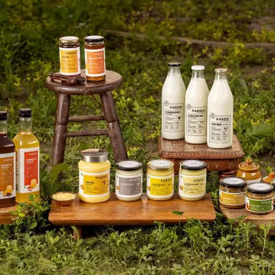 A complete product family shot for Barosi, featuring their range of ghee, oils, and milk in a rustic outdoor setting. This image communicates the brand's farm-fresh and natural ethos by placing the products right in the midst of greenery.