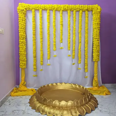A classic Haldi ceremony backdrop. This simple and elegant design features a frame draped in yellow fabric with hanging marigold garlands, paired with a large golden lotus-shaped bowl (urli).