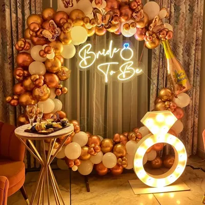 A glamorous "Bride To Be" setup perfect for a bachelorette party. This decoration features a circular balloon arch in white and chrome gold, a neon sign, butterfly accents, and a large light-up diamond ring prop.