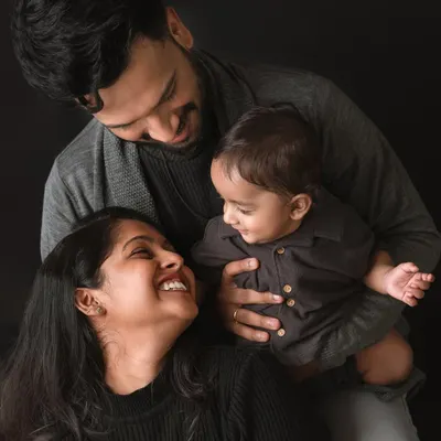 A classic family portrait that radiates warmth and connection. The dark, coordinated outfits create a timeless, fine-art feel, drawing all the attention to the loving expressions and smiles shared between the parents and their baby.