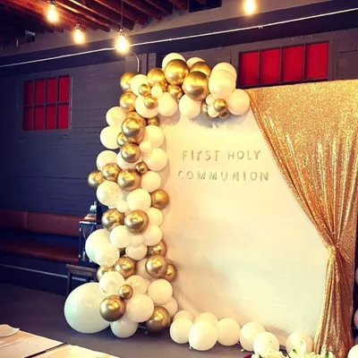 An elegant First Holy Communion decoration featuring a white and gold balloon arch against a white backdrop with a gold sequin curtain. This creates a sophisticated and celebratory setting for the milestone.