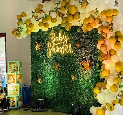 A stylish baby shower decoration featuring a lush green foliage wall with a 'Baby Shower' neon sign. The wall is framed by a white and yellow balloon arch with butterfly details.