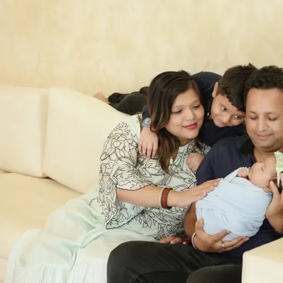 A candid and heartwarming lifestyle family photo. The family is relaxed on a sofa, with the older brother peeking over to get a closer look at his new baby sibling held by their father.