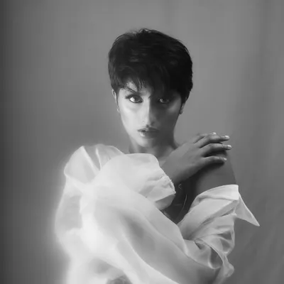 A soft-focus, ethereal black and white portrait of a woman with a pixie cut, wrapped in a sheer white blouse. The dreamy glow gives this studio headshot an artistic and gentle quality.