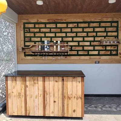 A complete view of a custom bar installation on a covered terrace. The setup includes a rustic wood-paneled counter with a dark top, paired with the signature bottle-shaped wall shelf and decorative paneling.