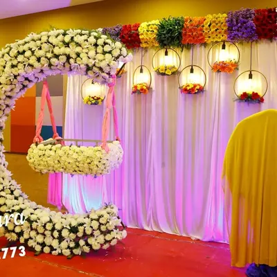 A stunning cradle ceremony setup featuring a crescent-shaped arch made of white flowers. The hanging cradle, also adorned with flowers, creates a beautiful focal point, while colorful lanterns add a festive touch.