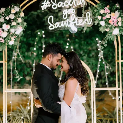 A romantic moment for the newly engaged couple, framed by a modern golden arch decorated with pastel flowers and a custom neon sign. The fairy lights and lush green background add a touch of magic.