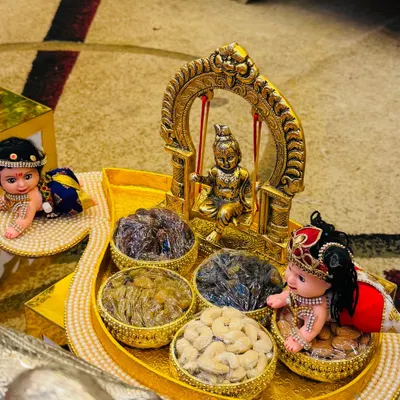 A charming dry fruit platter for a baby naming ceremony. The centerpiece is a golden swing with a baby Krishna idol, surrounded by bowls of cashews and raisins and cute baby dolls.