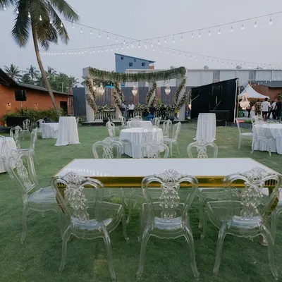 An elegant outdoor reception setup on a lush green lawn. The combination of modern ghost chairs, classic chandeliers, and string lights creates a sophisticated yet relaxed atmosphere.