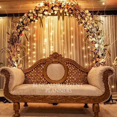 I designed this elegant stage for a wedding reception, featuring a classic carved sofa set against a backdrop of warm fairy lights. The setup is framed by a lush floral arch, creating a beautiful and timeless focal point for the couple.