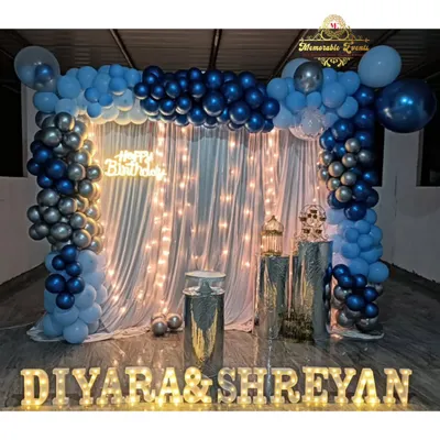 A beautiful birthday setup with a blue and silver balloon arch over a backdrop of sheer curtains and fairy lights. The names are highlighted with marquee letters.