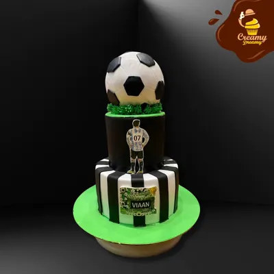 A two-tier football theme cake perfect for a young sports fan. The design includes a striped bottom tier, a player cutout, and a realistic fondant football on top.