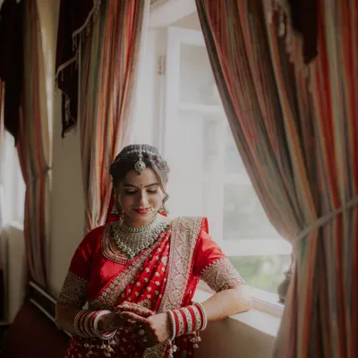 An elegant bridal portrait of the bride in her red saree, captured in beautiful natural light by a window. These quiet, reflective moments before the ceremony make for timeless photos.