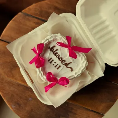 A beautiful heart-shaped bento cake with the word "blessed" and the angel number "11:11" piped on top. The delicate pink satin bows add a sweet, romantic touch.