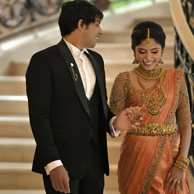 A classic and elegant look for the groom. This perfectly tailored black tuxedo with a satin lapel is a timeless choice that stands proudly next to the bride's traditional orange Kanjivaram saree.
