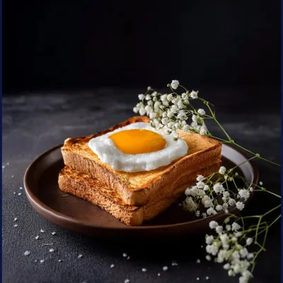 A perfect sunny-side-up egg on golden toast, styled with delicate baby's breath flowers against a dark, textured background. This shot balances rustic simplicity with elegance, ideal for a cafe's breakfast menu.